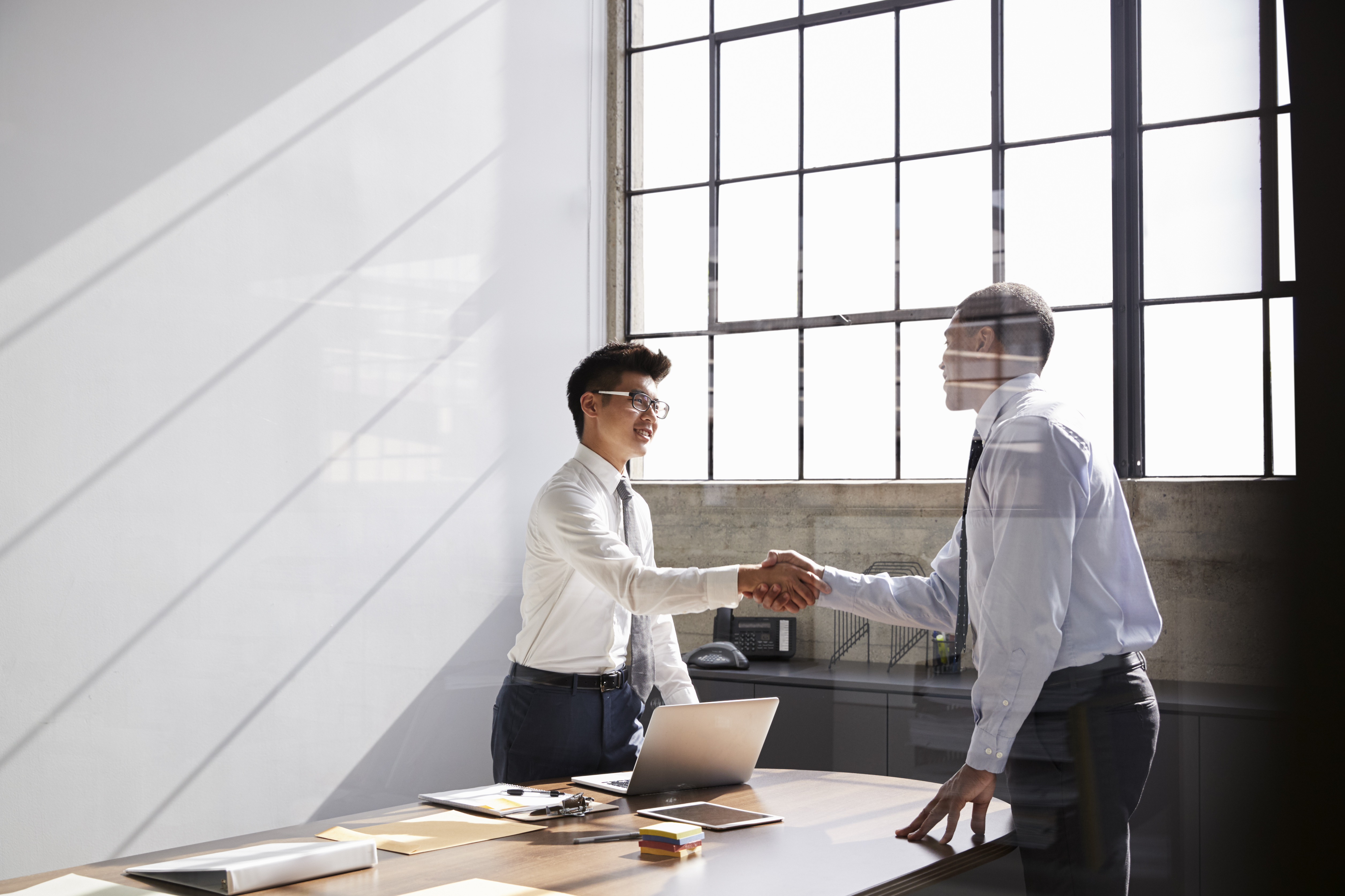 Two men in formal attire shake hands across a desk in a bright office. Sunlight streams through large windows, creating a professional and positive atmosphere.