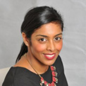 Smiling person with dark hair in a black outfit and necklace against a gray background.