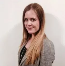 Woman with long hair in a grey blazer standing against a white wall.