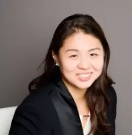Smiling person in a black blazer, seated against a gray background.
