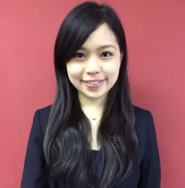 A woman with long dark hair wearing a black jacket smiles in front of a red background.