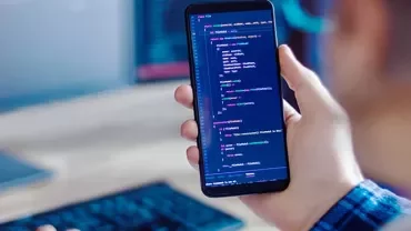 Person holding a smartphone displaying colorful code, with a blurred computer screen in the background.
