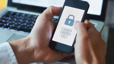 Person holding smartphone with security lock on screen, laptop in background.