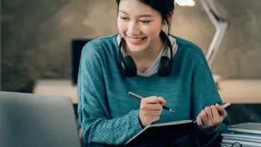 Woman smiling, taking notes at a desk with a laptop and headphones.
