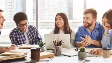Diverse group of students laughing and studying together with laptops and coffee in a modern setting.