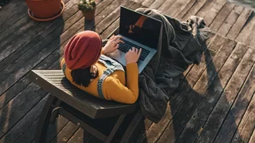 Person in a red hat using a laptop on a wooden deck near water.