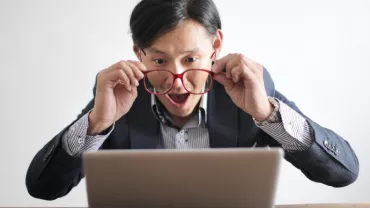 An Asian, white-collar professional holding out his glasses for a better look at his laptop screen.