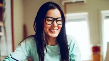Smiling person with glasses working at a laptop in a cozy room.