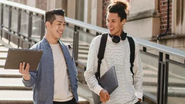 Two students walking down stairs, holding laptops and smiling.