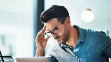 Man in glasses working on a laptop, looking focused in casual office setting.