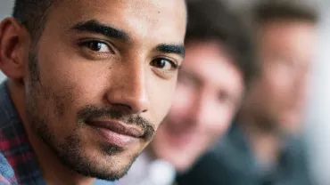 Man in a plaid shirt smiling with colleagues in a blurred background.