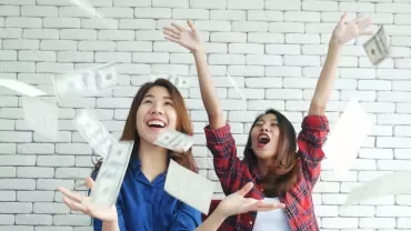 Two women happily throwing money in the air against a white brick wall.