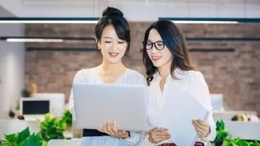 Two women in an office collaborate over a laptop, discussing career growth opportunities and talent retention strategies.