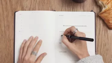 Person writing in a notebook with a pen, croissant on table.