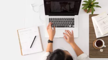 Overhead view of person working on a laptop with a planner, plant, and cup of coffee on the desk.
