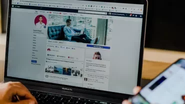Person using a laptop, browsing a social media profile, while holding a smartphone.