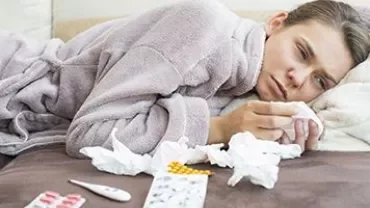 Person in a robe lying on a couch surrounded by tissues and medication.
