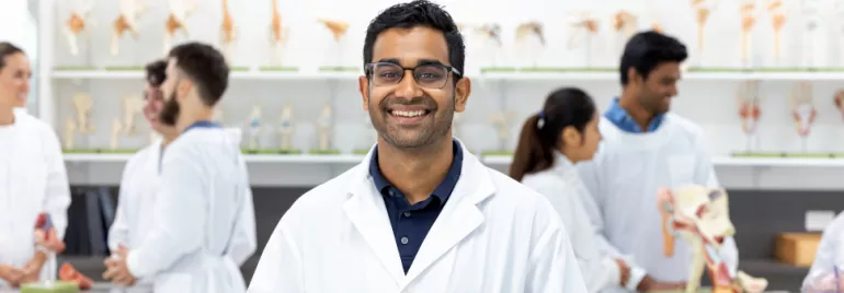 A smiling lab technician in a white coat stands in a science classroom with colleagues working in the background.