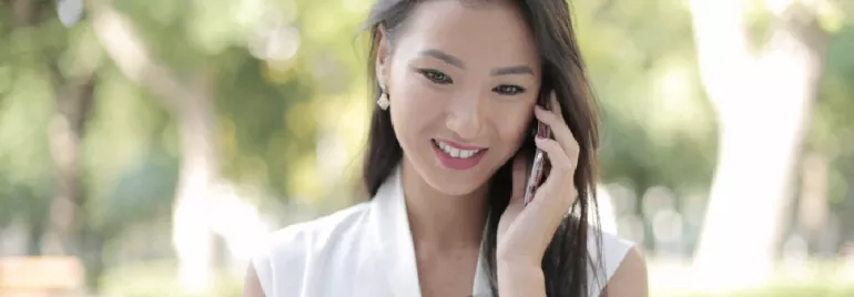 Asian female woman in her 30s, smiling while talking on a mobile phone and looking at her computer screen