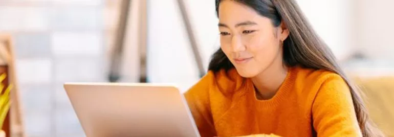 Woman in orange sweater working on a laptop at a desk.
