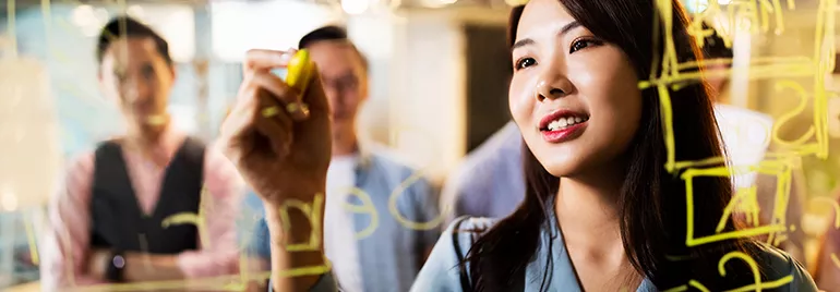 Woman writing on transparent board with yellow marker, people in background.