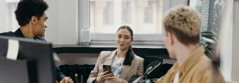 Three people having a meeting in an office, two are sitting and one is holding a phone.