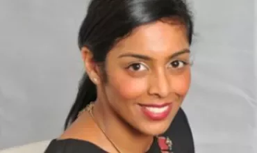 Smiling woman with dark hair and red necklace against a grey background.