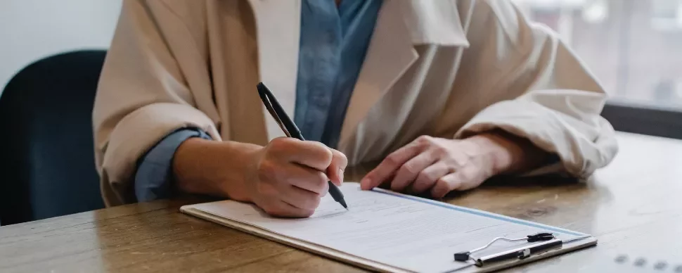 Person signing a document on a clipboard at a wooden table.