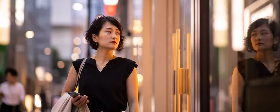 Woman walking past a storefront, holding a bag and phone.
