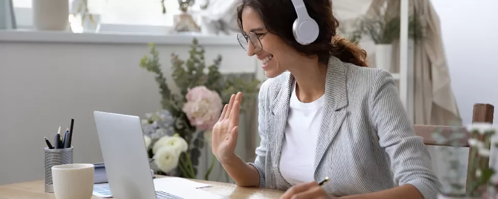 Woman with headphones using laptop, smiling and waving.