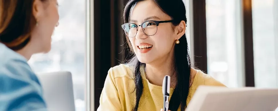 Two women having a conversation at a table, one holding a pen and smiling.