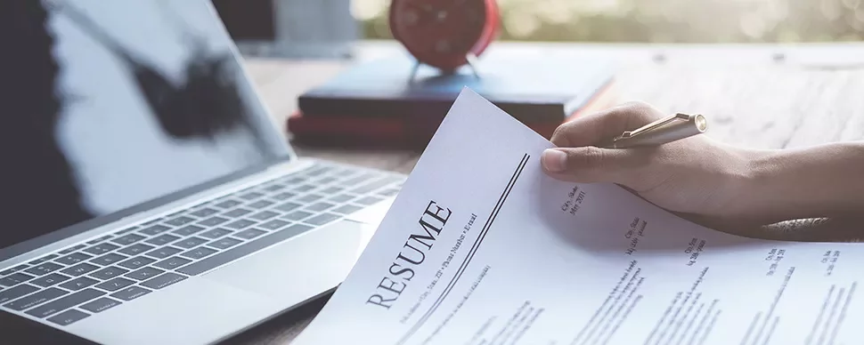 Person holding a resume next to an open laptop on a desk.