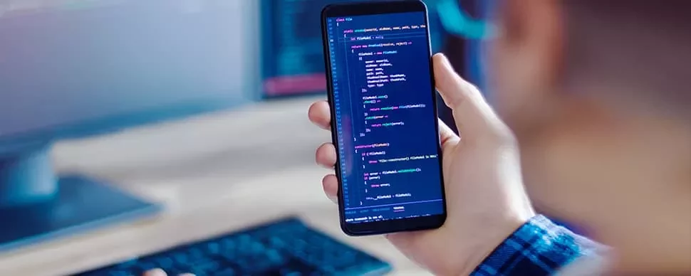 Person holding a smartphone displaying colorful code, with a blurred computer screen in the background.