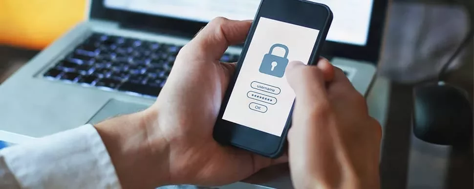 Person holding smartphone with security lock on screen, laptop in background.