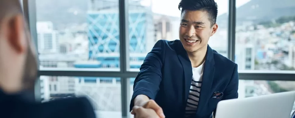 Smiling man in a suit shaking hands across a desk in an office.