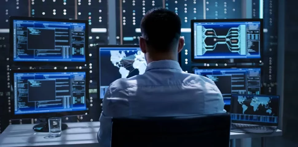 Person working in a data center, using multiple monitors displaying code and world maps.