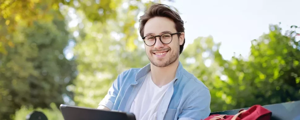 Man with glasses smiling while sitting outside with greenery in the background.