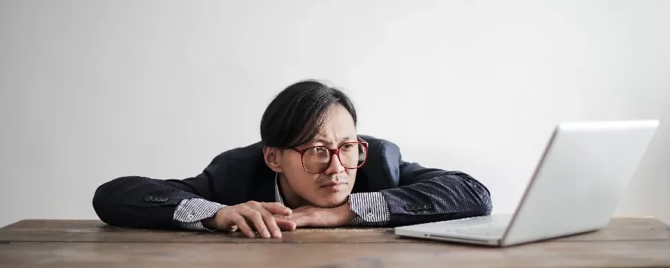 Man in suit and glasses staring thoughtfully at an open laptop on a wooden table.