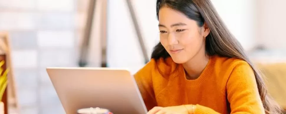 Woman in orange sweater working on a laptop at a desk.