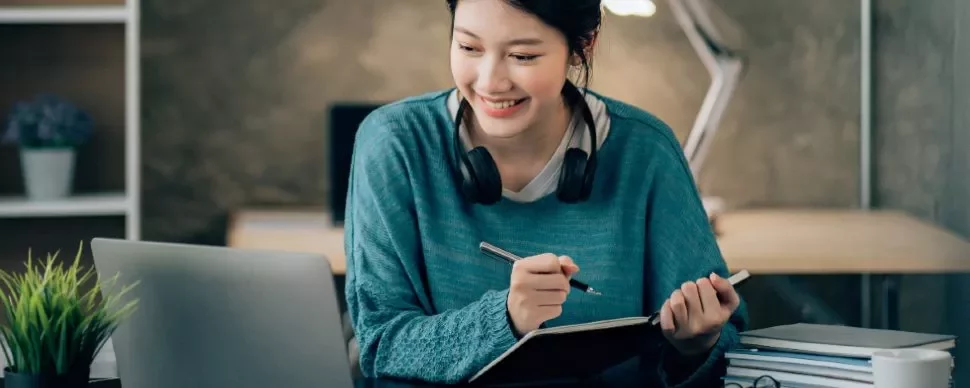 Woman smiling, taking notes at a desk with a laptop and headphones.