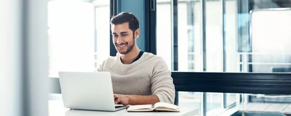 Man smiling while using a laptop at a desk with a notebook open.