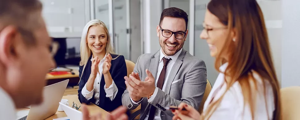 A group of business professionals sitting at a table, laughing and clapping during a meeting.