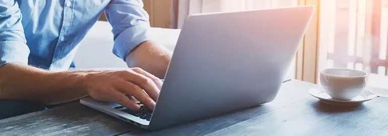 Person typing on a laptop at a wooden table with a cup of coffee nearby.