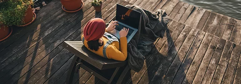 Person in a red hat using a laptop on a wooden deck near water.