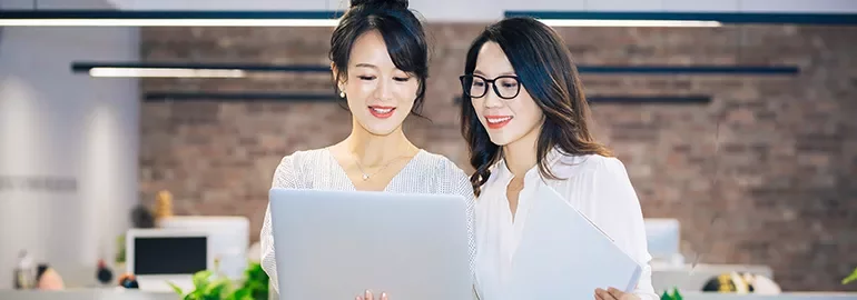 Two women smiling and looking at a laptop in an office environment.