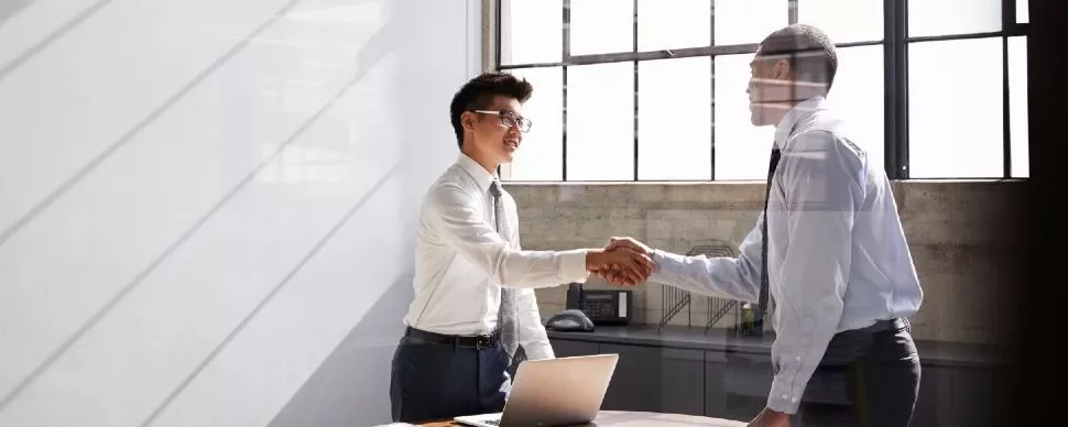 Two men in an office shaking hands by a desk with a laptop.