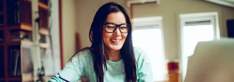 Smiling person with glasses working at a laptop in a cozy room.