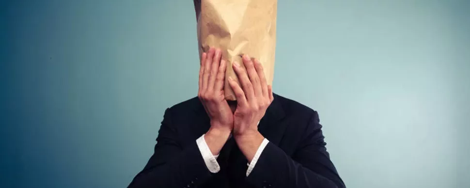 Person in a suit with a paper bag over their head against a blue background.