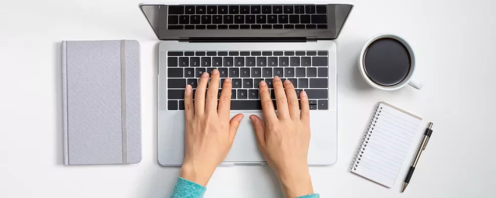 Hands typing on a laptop next to a notebook and coffee cup.