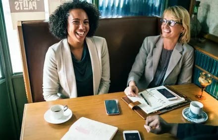 Two women smiling and chatting at a cafe table with papers and coffee.